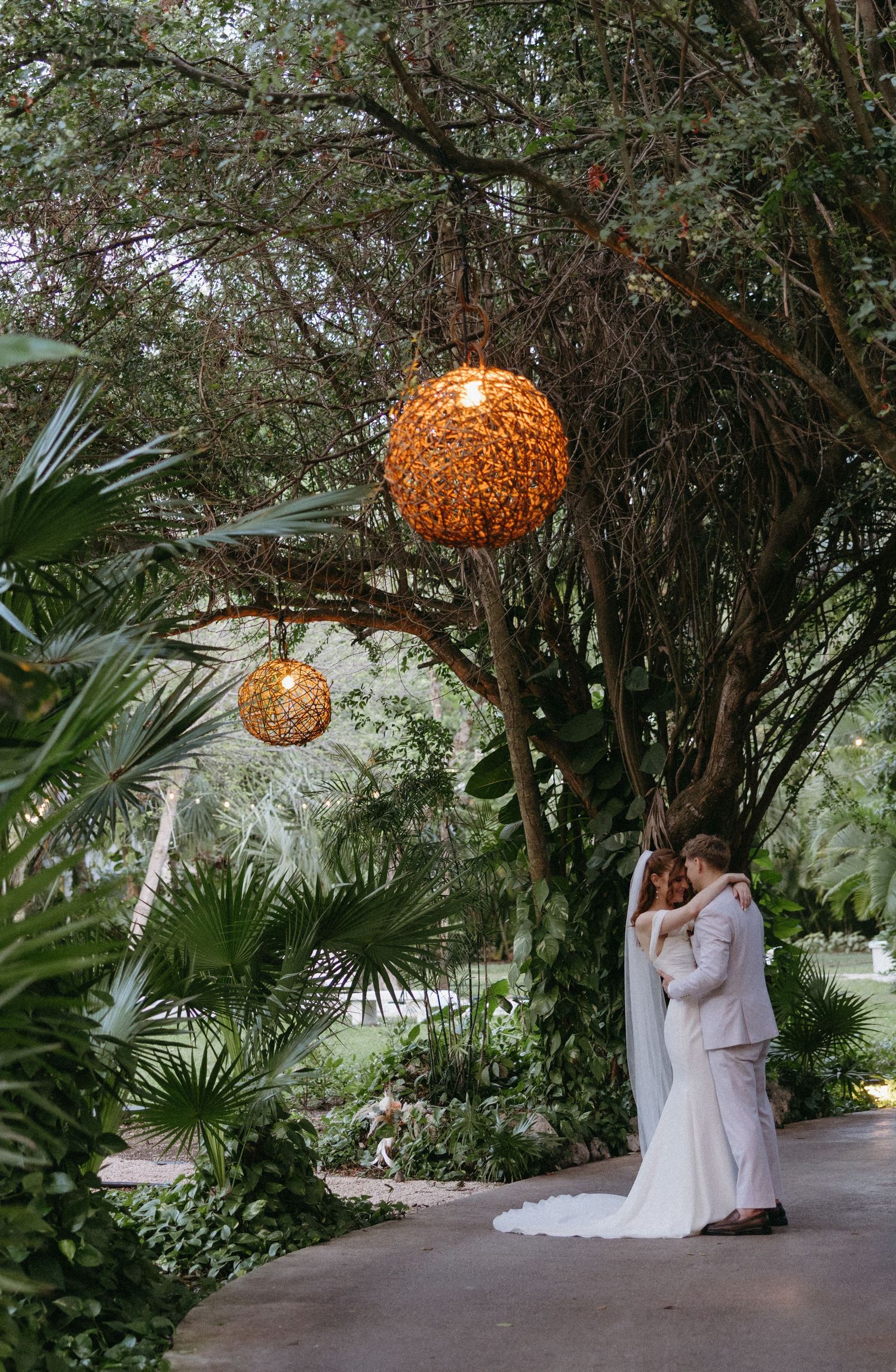 Wedding couple embraces under glowing globe lanterns in lush tropical garden setting.
