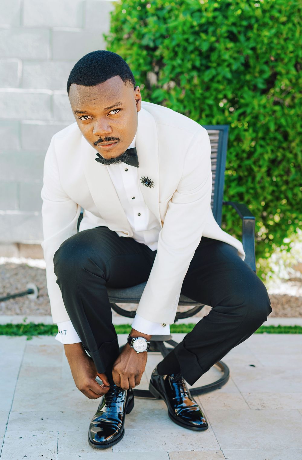 Groom in white tuxedo jacket tying black patent leather shoes outdoors.