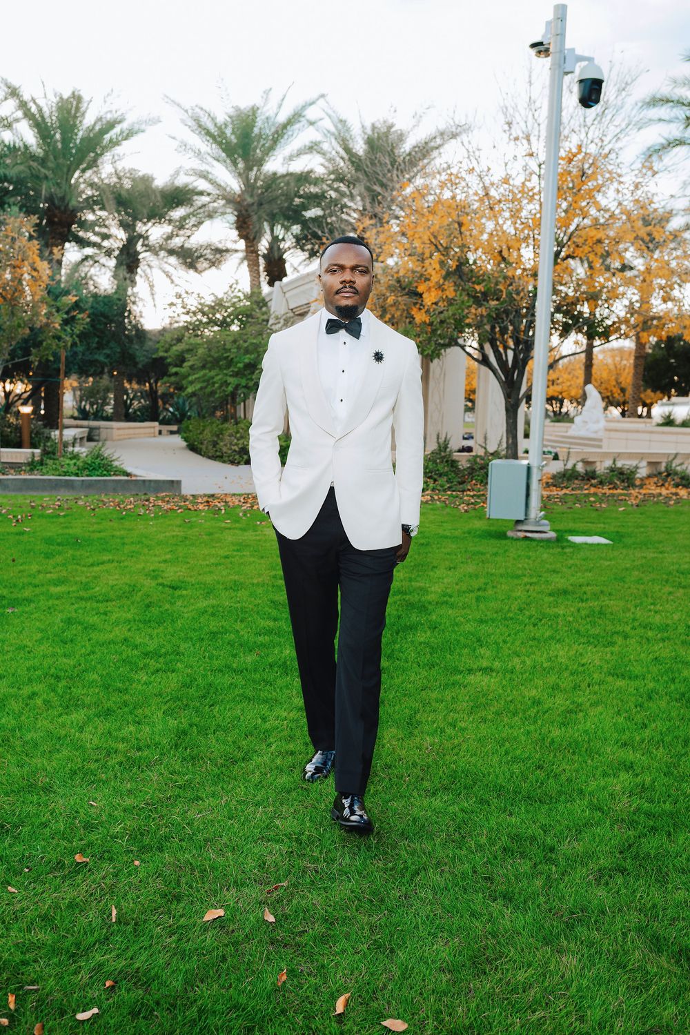 A well-dressed Black man in a white tuxedo jacket and black pants walks across green grass near palm trees.