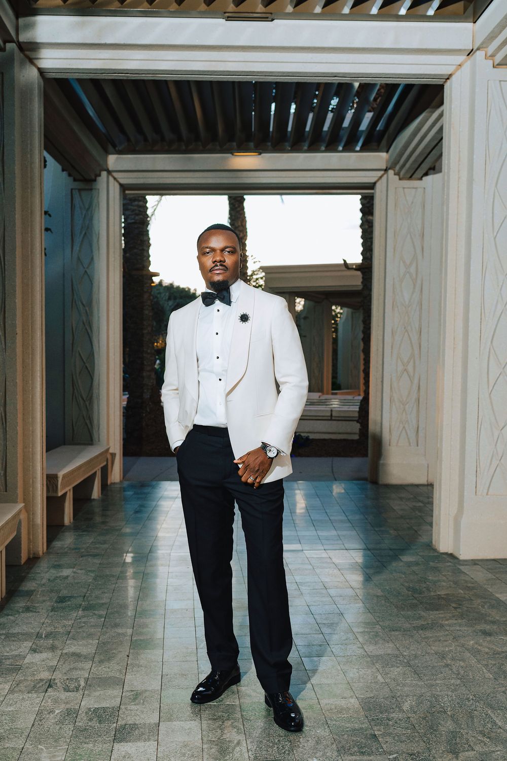 A well-dressed man in a white tuxedo jacket and black bow tie stands confidently in an elegant outdoor venue.