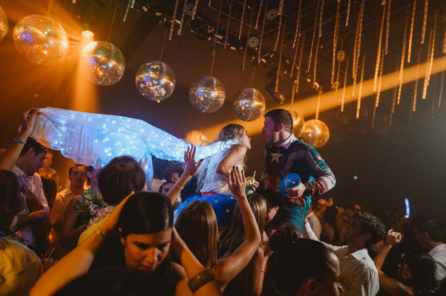 Bride and groom lifted by cheering guests under decorative globe lights at wedding reception.