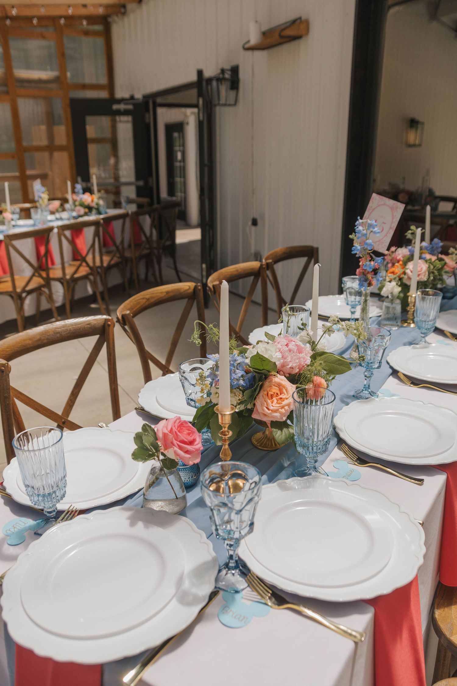 Elegant wedding reception table with white plates, blue glasses, pink floral centerpiece, candles, and wooden cross-back chairs.
