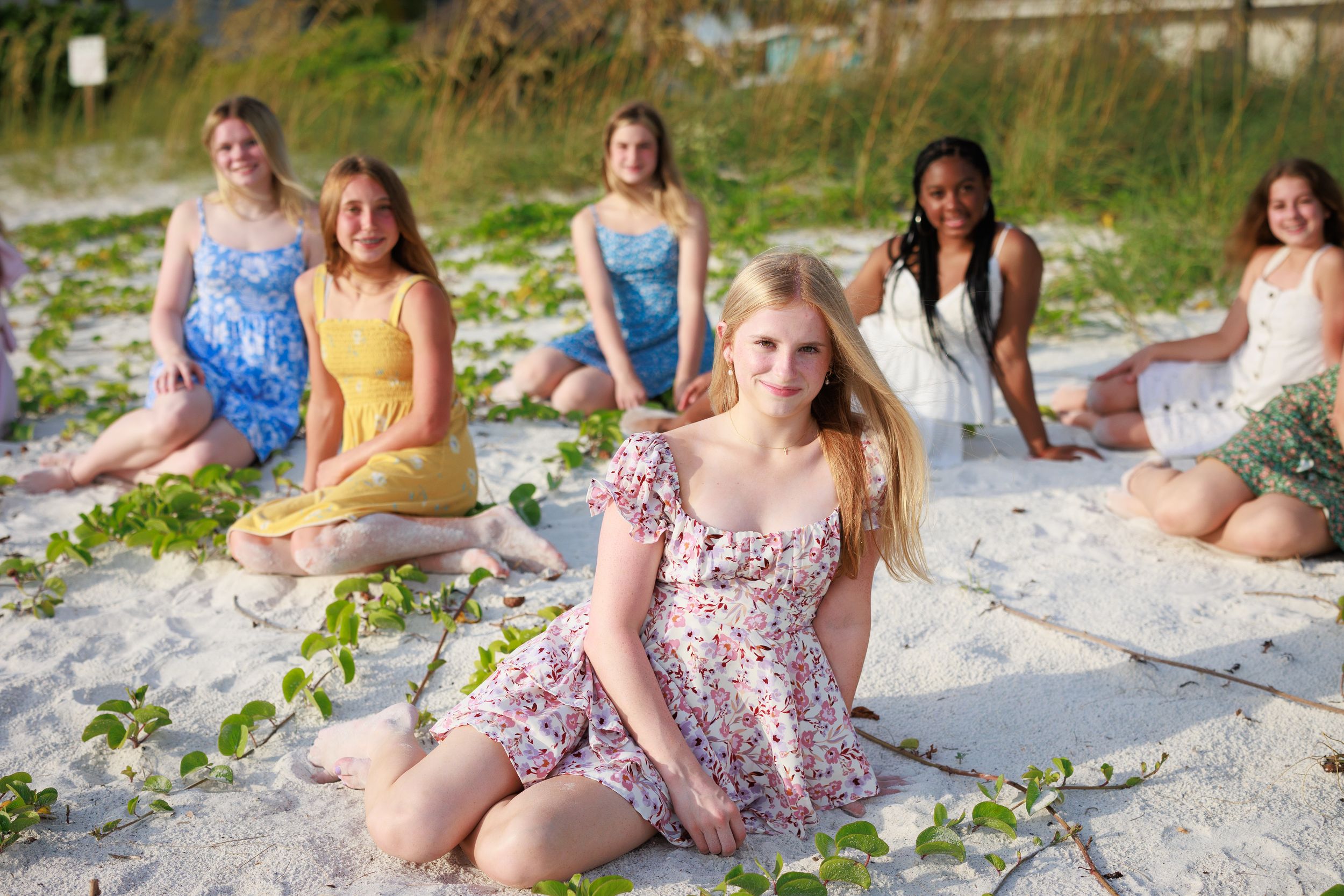 Group of young girls in colorful dresses posing together on a sandy beach with green vines.