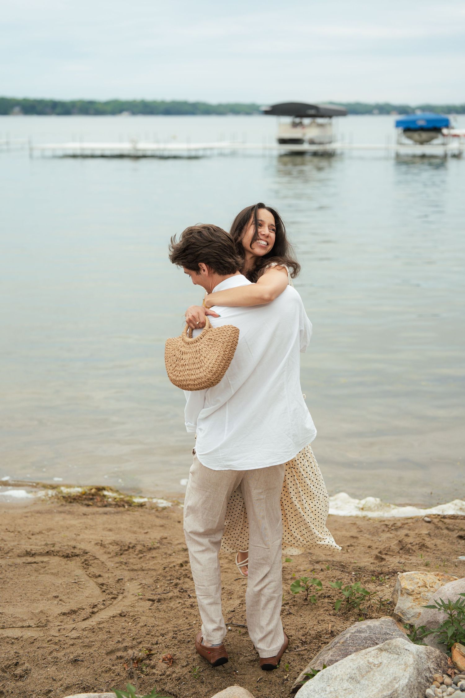 Couple embracing and smiling by a lakeside dock, wearing casual neutral tones, with sandy shore and boats in background.