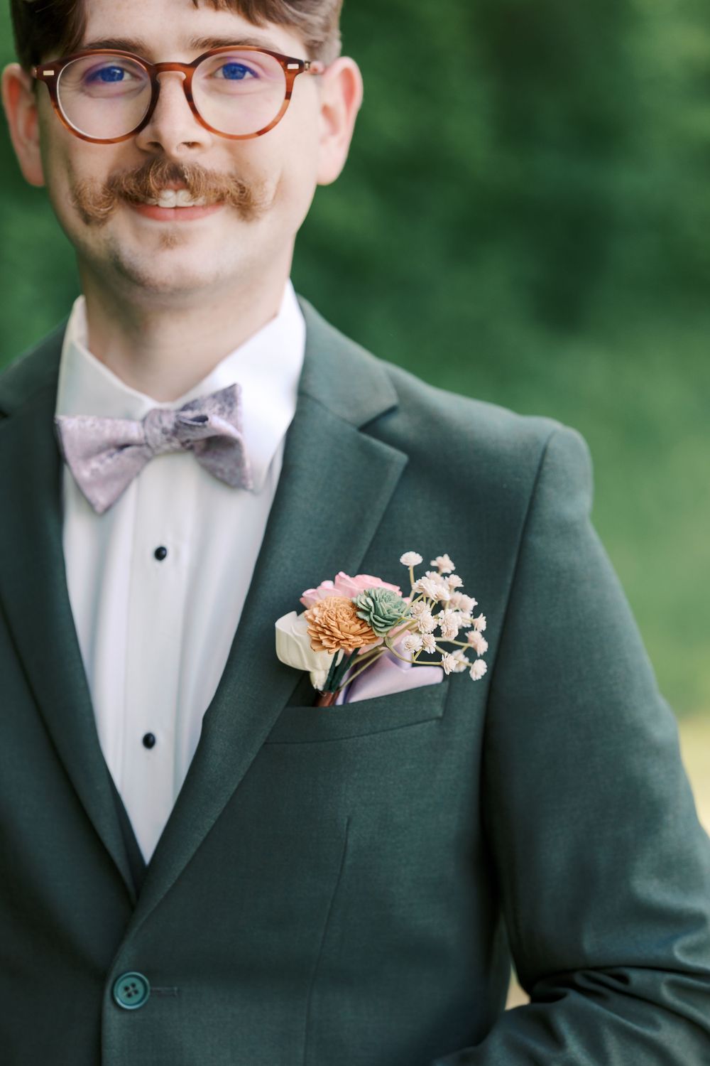 Smiling groom in green suit with floral boutonniere and lavender bow tie wearing round glasses outdoors.