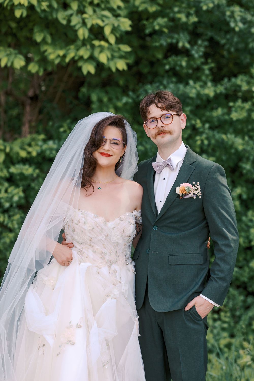 Bride and groom pose together outdoors, she in floral off-shoulder gown with veil, he in dark green tuxedo.