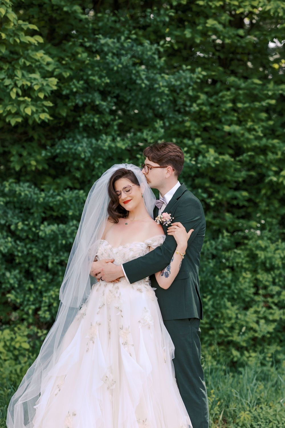 Groom in green suit tenderly embraces bride in floral ballgown and veil against lush green foliage backdrop.