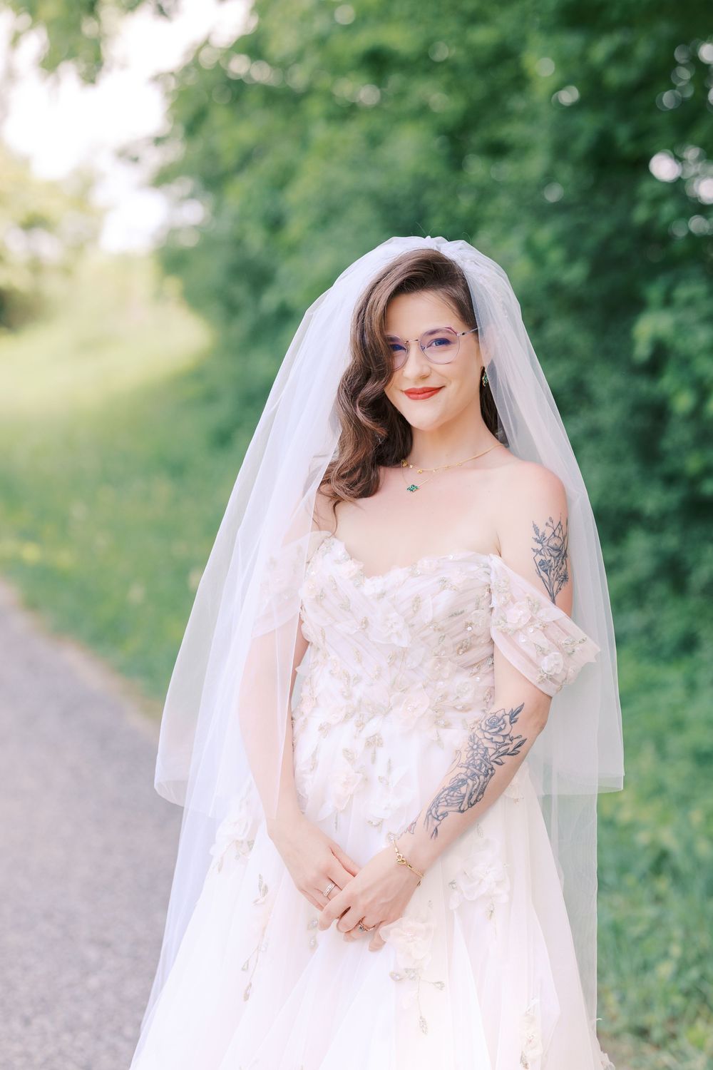 Tattooed bride in off-shoulder floral gown and veil smiles softly on sunlit tree-lined path outdoors.