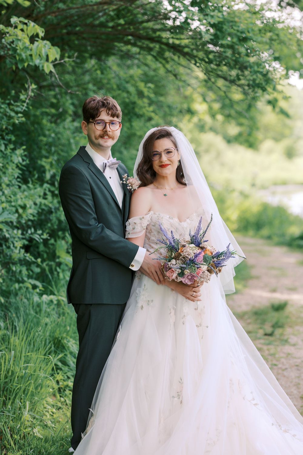 Bride and groom pose together outdoors among lush green trees, she holds a colorful wildflower bouquet in a white gown.