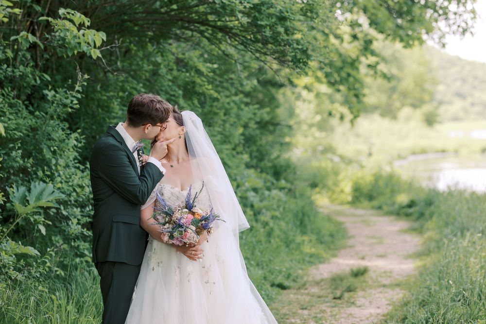 Bride and groom share a tender moment on a lush green nature path holding a wildflower bouquet.