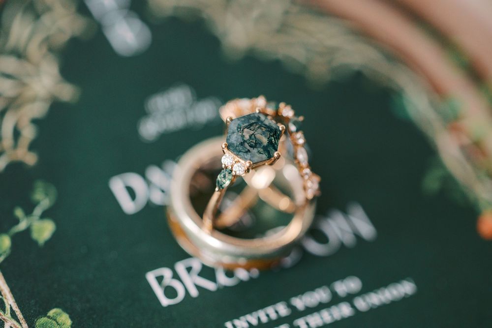 Gold engagement ring with raw dark gemstone resting on green wedding invitation suite.