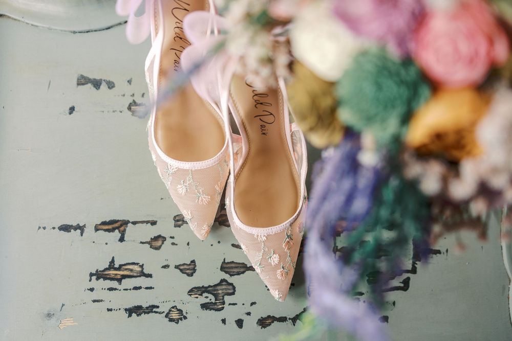Elegant blush bridal heels with floral embellishments displayed beside a colorful wedding bouquet on a vintage surface.