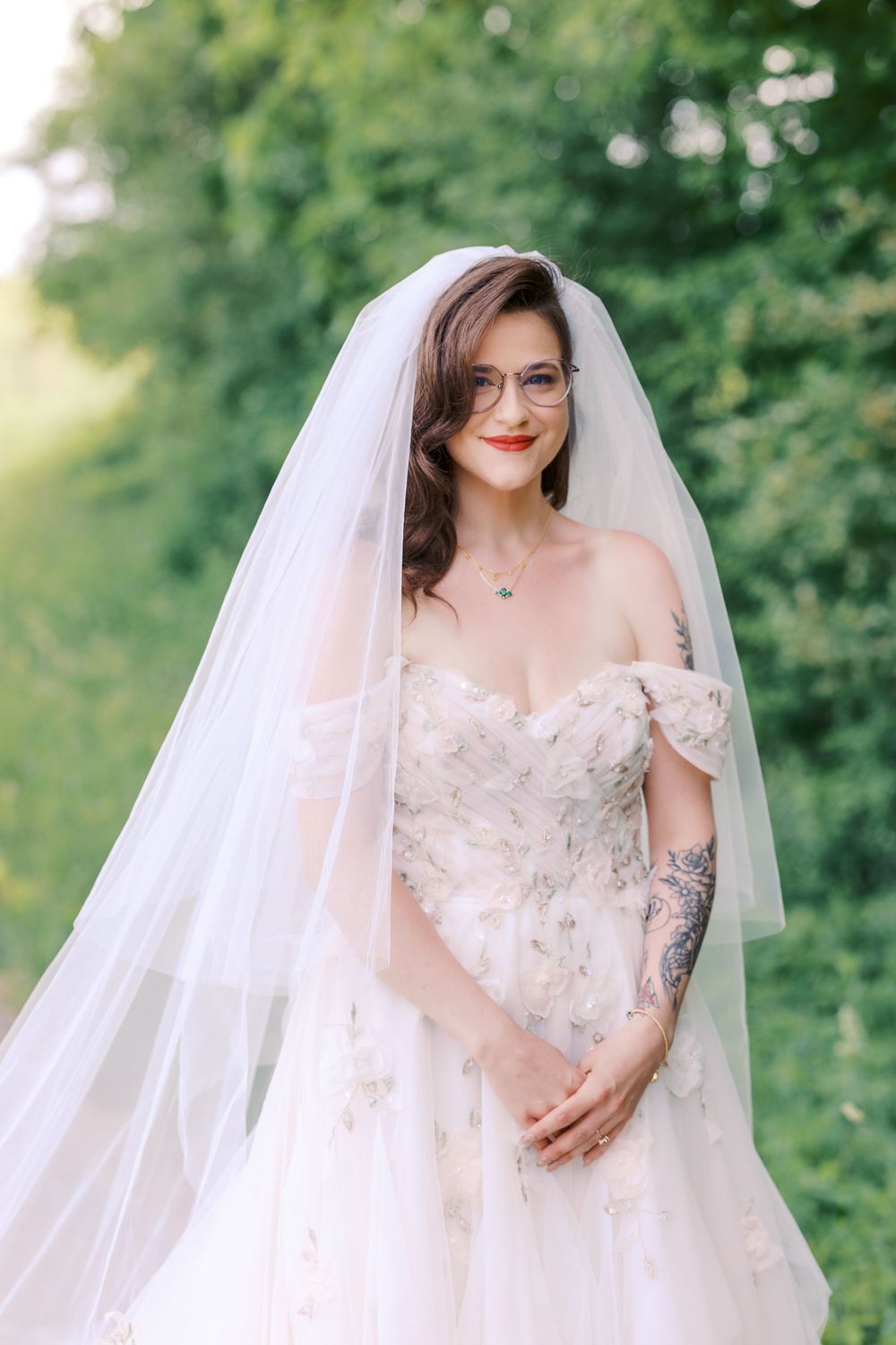Bride in off-shoulder floral wedding gown with veil and glasses smiling outdoors among green trees.