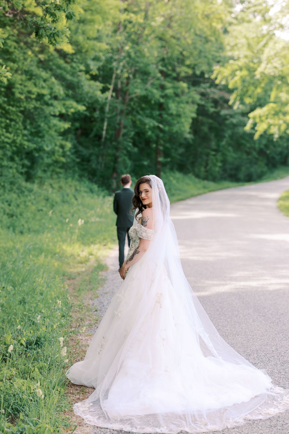 Bride in flowing white ballgown and veil walks with groom on tree-lined path in lush green outdoor setting.
