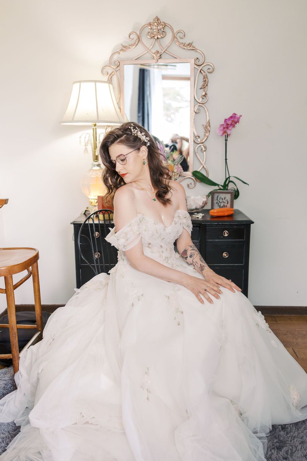 Bride in off-shoulder floral ballgown sits before vintage mirror with lamp and orchids in elegant bridal suite.