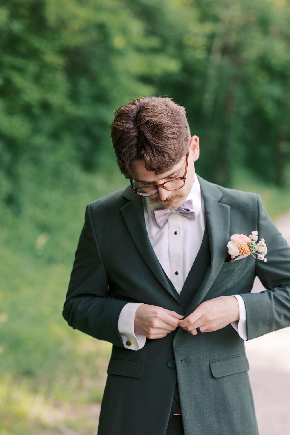 Groom in dark green tuxedo adjusting jacket button outdoors with lush green trees in background.