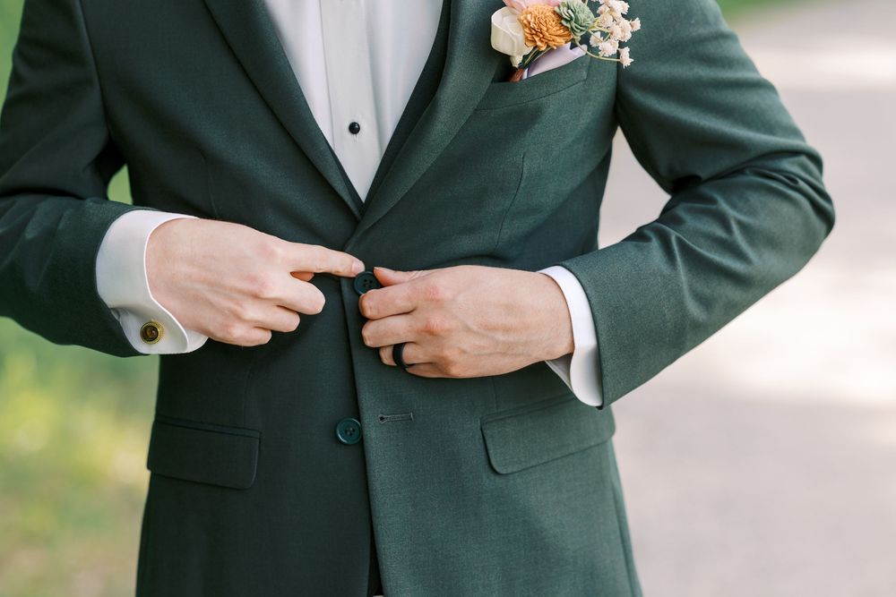 Groom in dark green suit adjusting jacket buttons, wearing cufflinks and floral boutonniere at wedding.