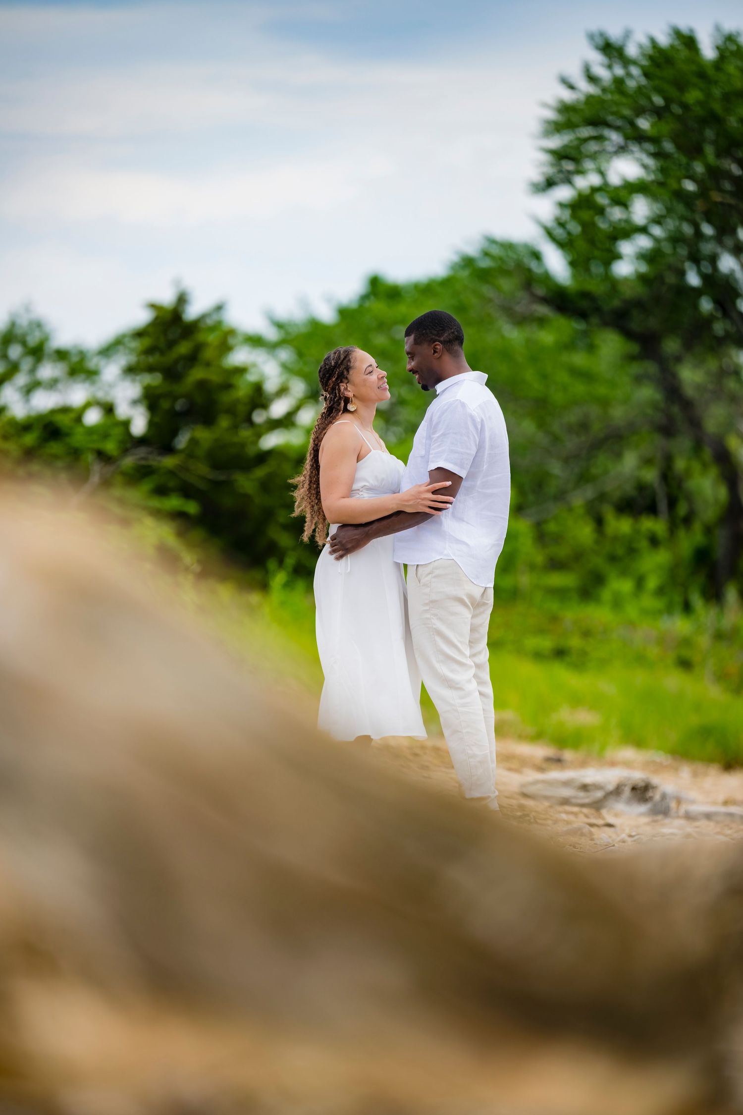 Interracial couple in white outfits embracing outdoors on sandy ground with lush green trees in background.