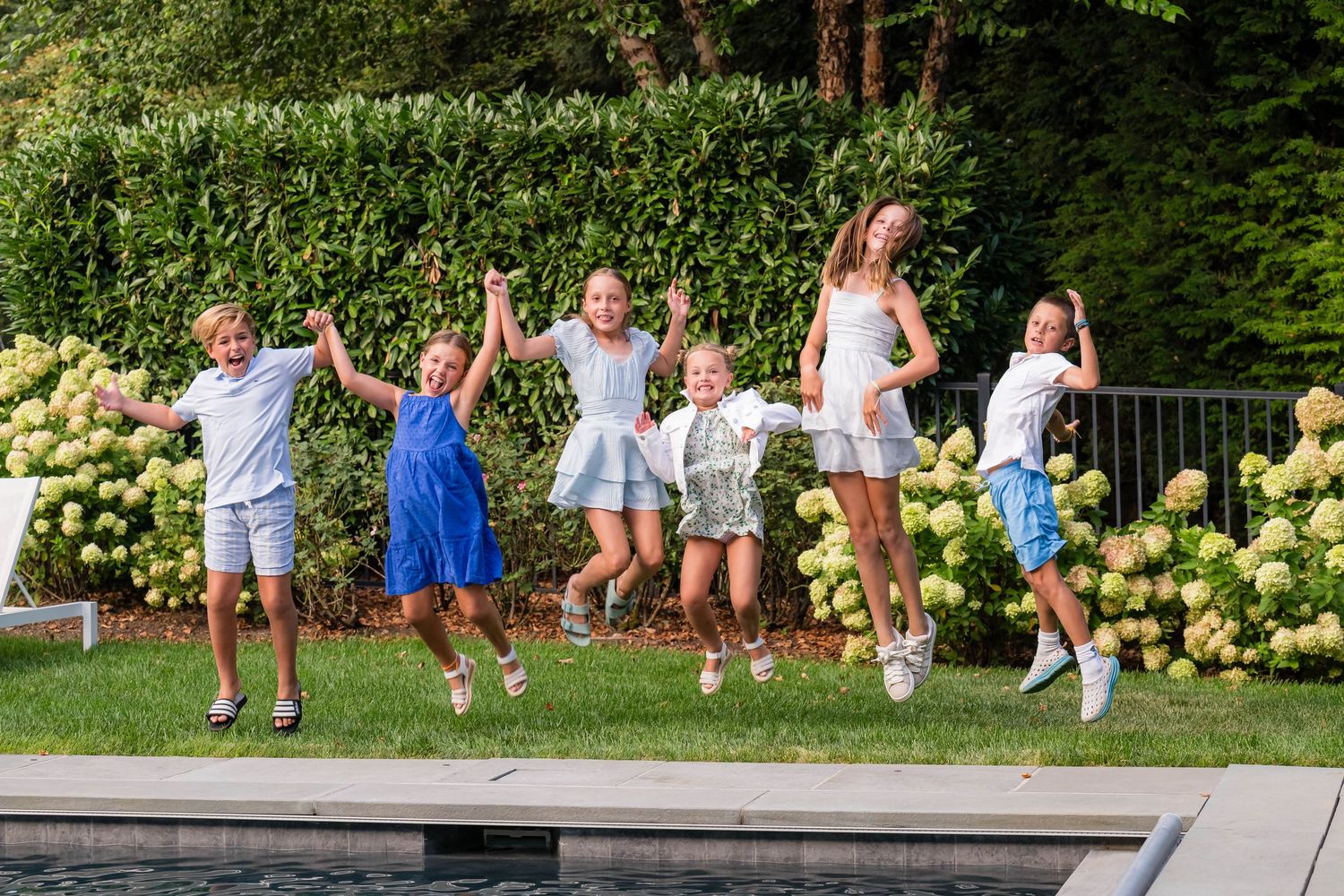 Six children joyfully jumping in unison outdoors near a pool, surrounded by lush green hedges and blooming hydrangeas.