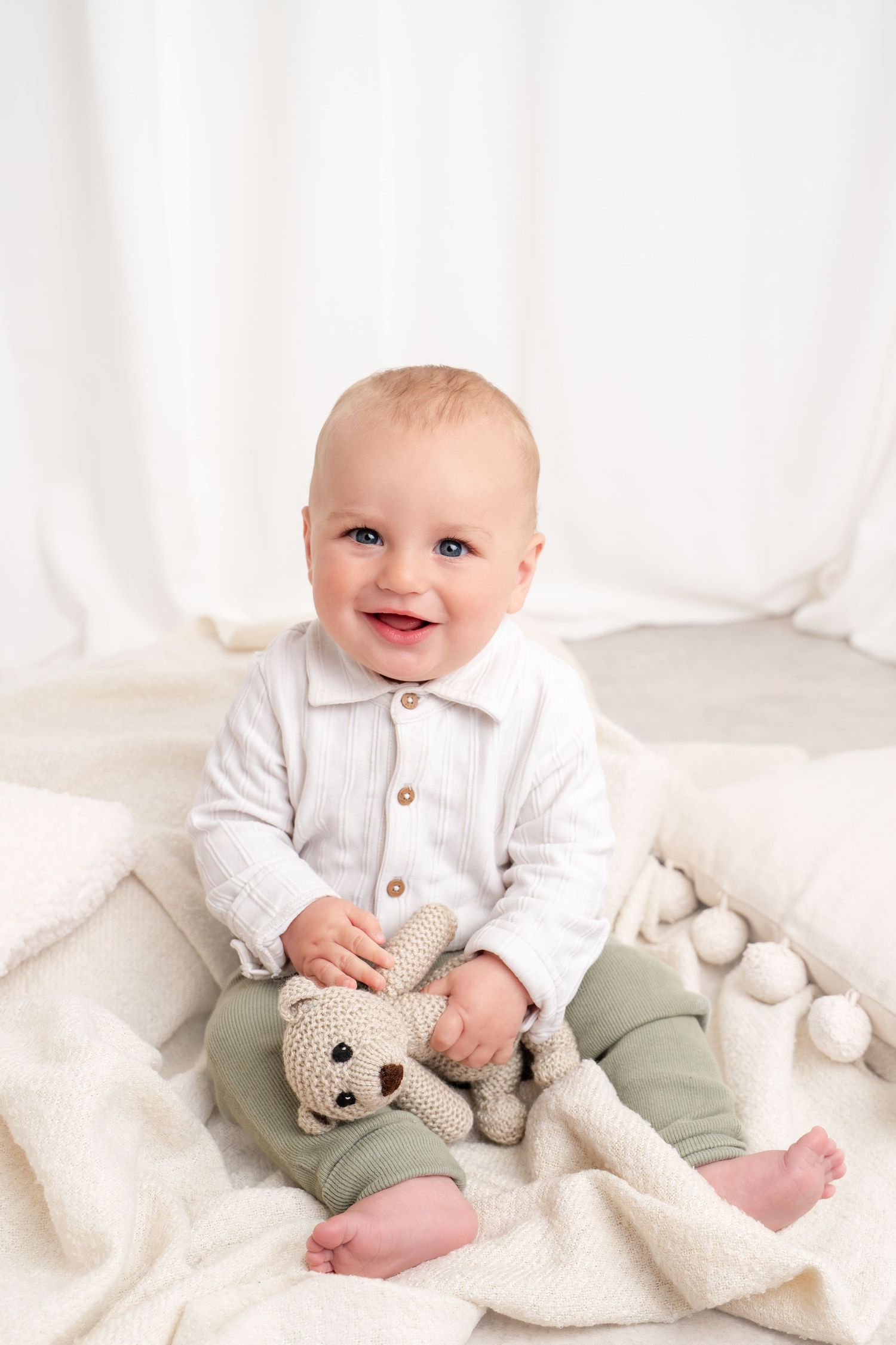 Smiling baby in white outfit holds knit teddy bear while sitting on fluffy white blanket.