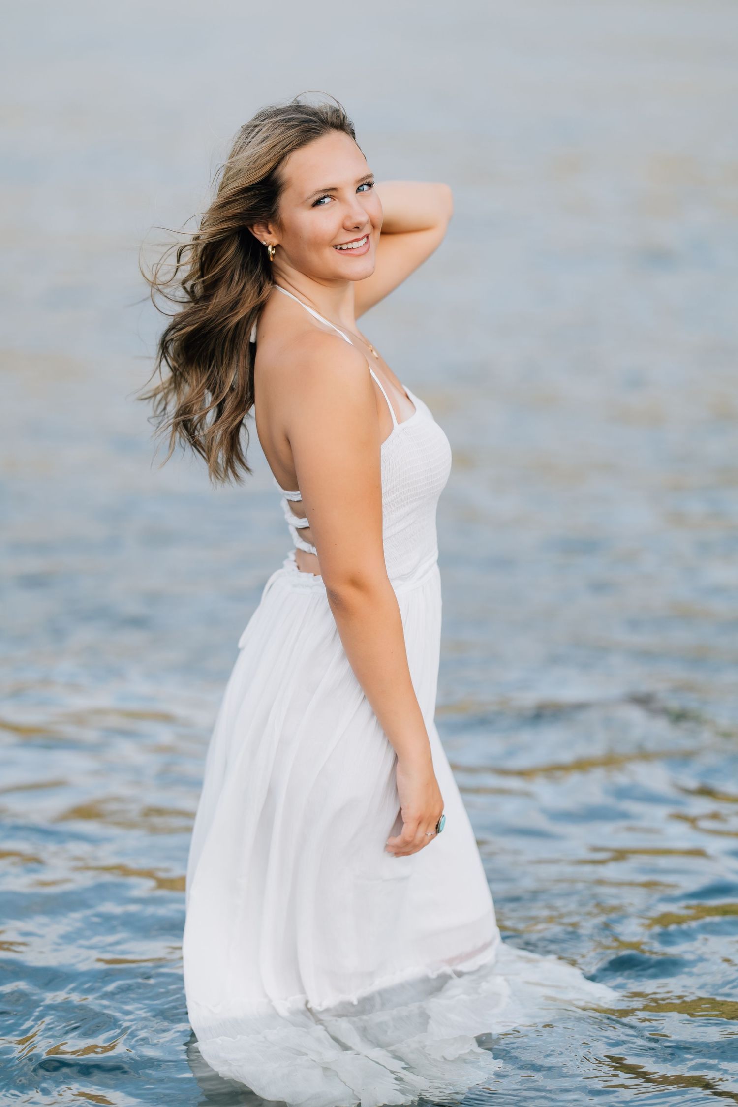 Young woman in white halter dress standing in shallow ocean water, smiling over shoulder with wind-blown hair.