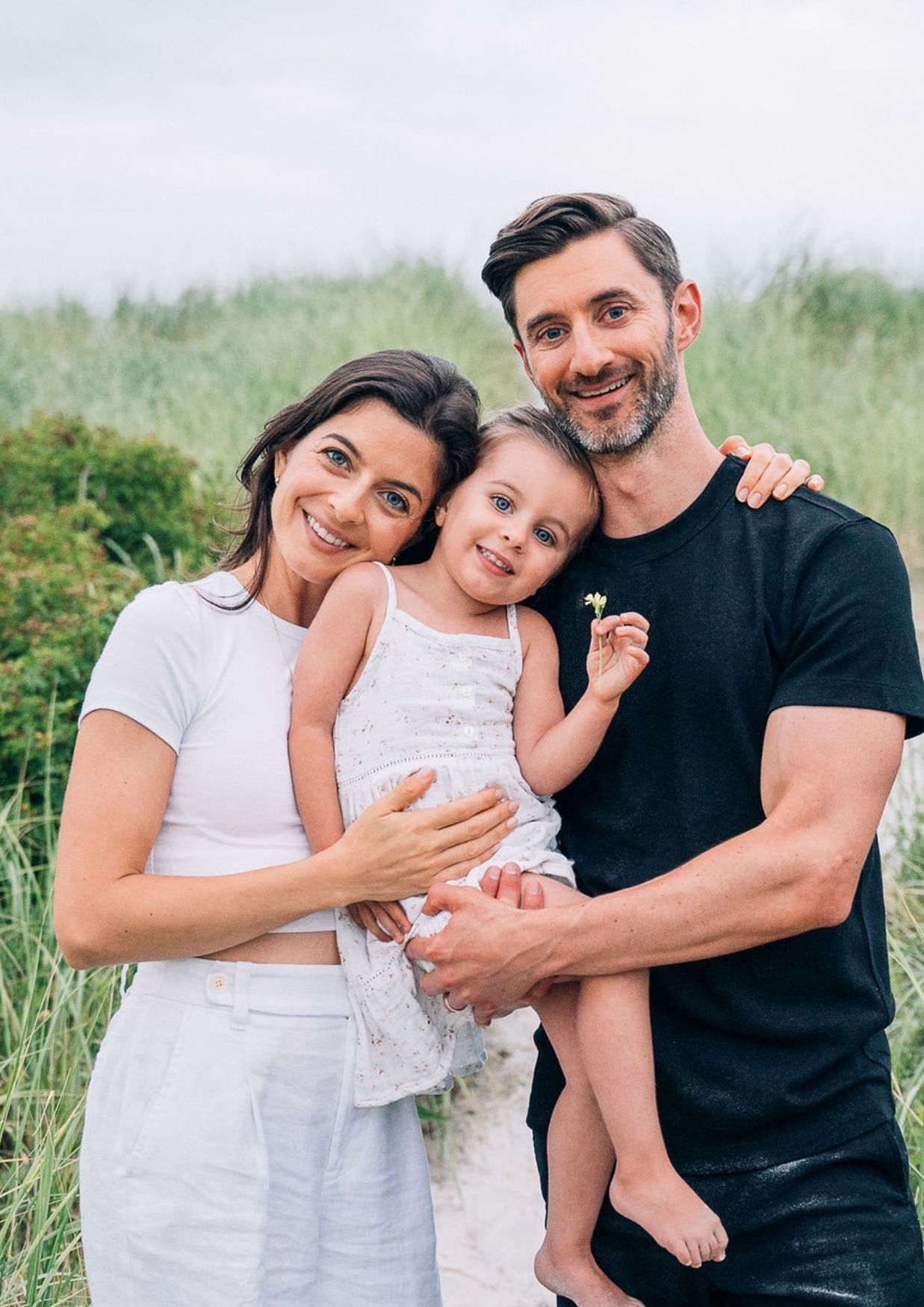 Happy family of three posing outdoors near beach grass, parents holding young daughter in white dress.