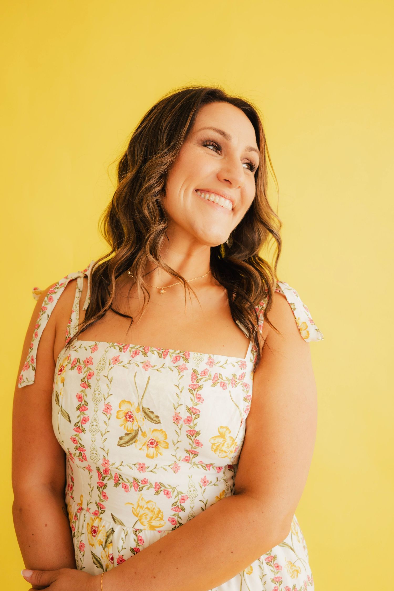 Smiling woman in floral sundress poses against bright yellow background with wavy brown hair.