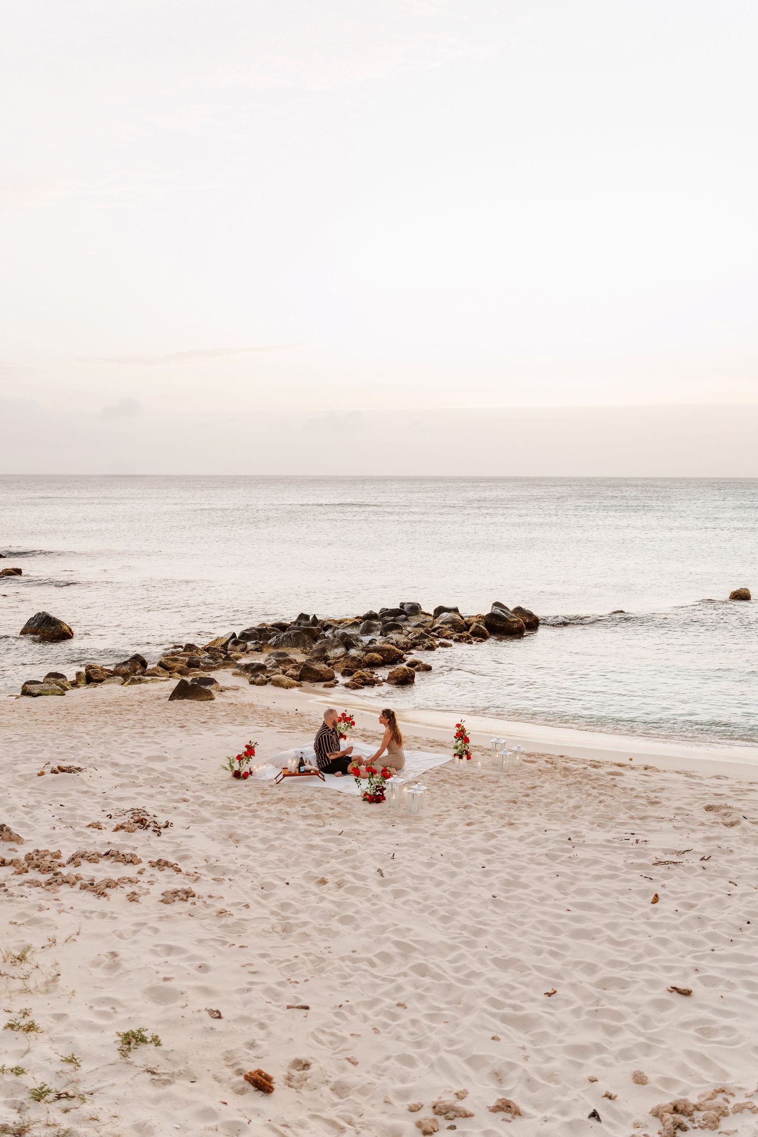 Serene white sand beach with calm blue ocean waters and a sailboat anchored near rocky shoreline under soft sky.