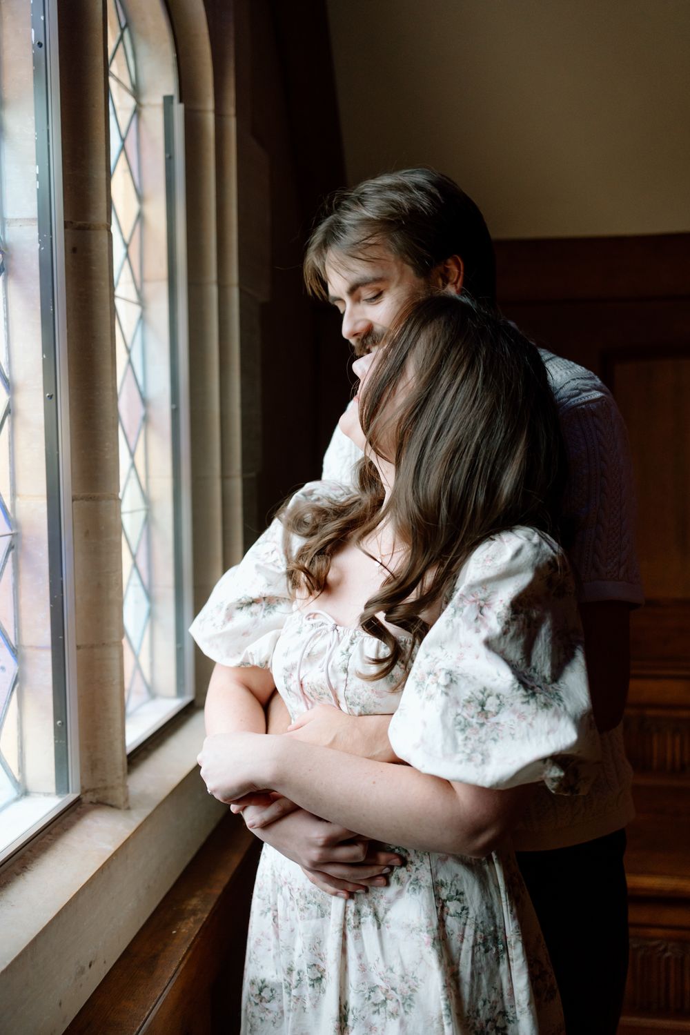 Man tenderly hugging woman from behind near a stained glass window in a warm, intimate indoor setting.