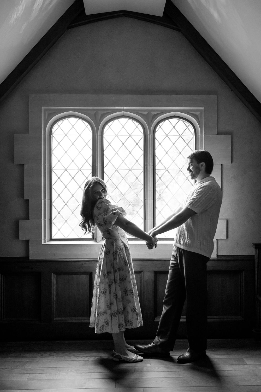 Romantic black and white silhouette of couple holding hands before arched leaded glass windows indoors.