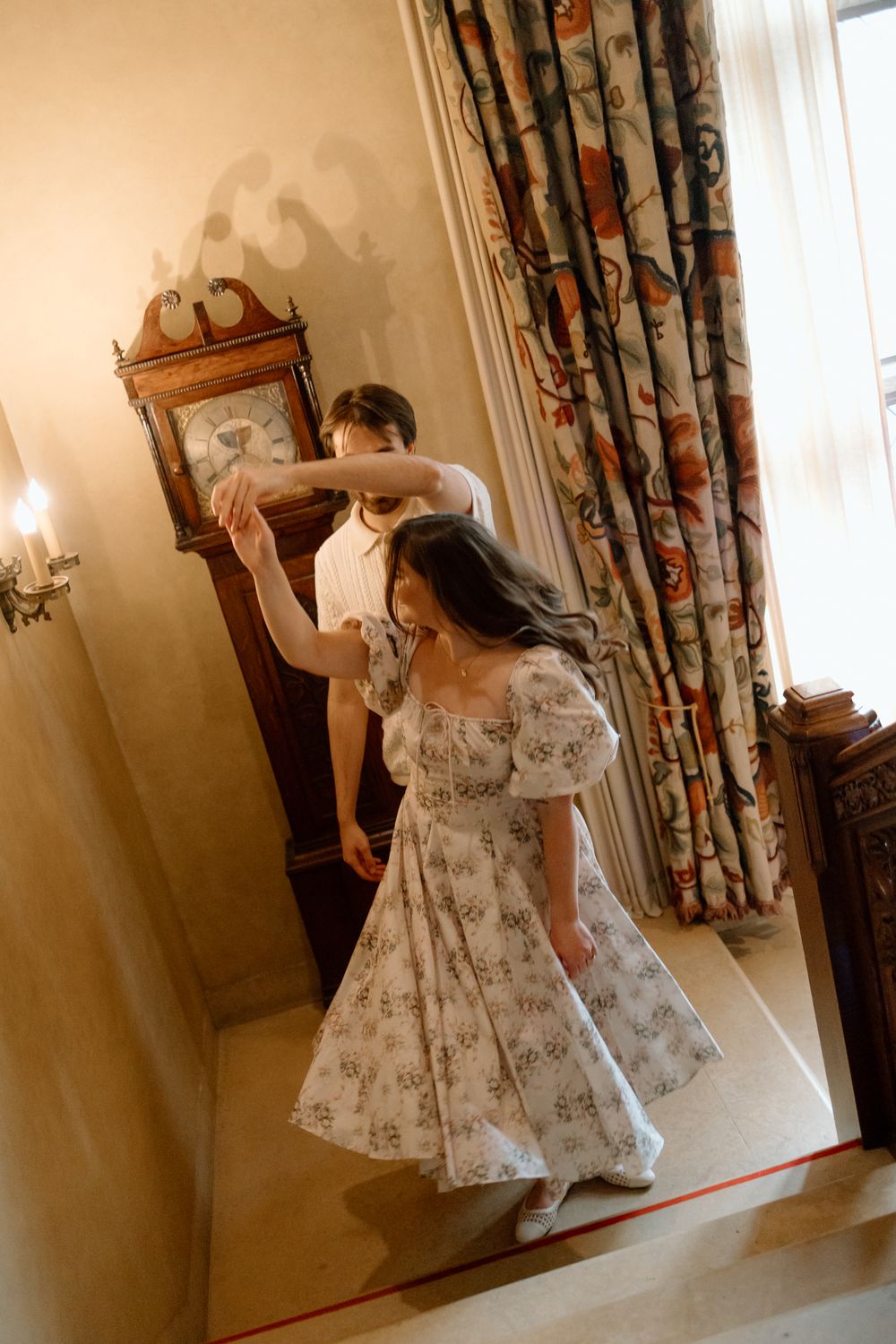 Couple dancing near an antique grandfather clock in a warmly lit elegant room with floral curtains.