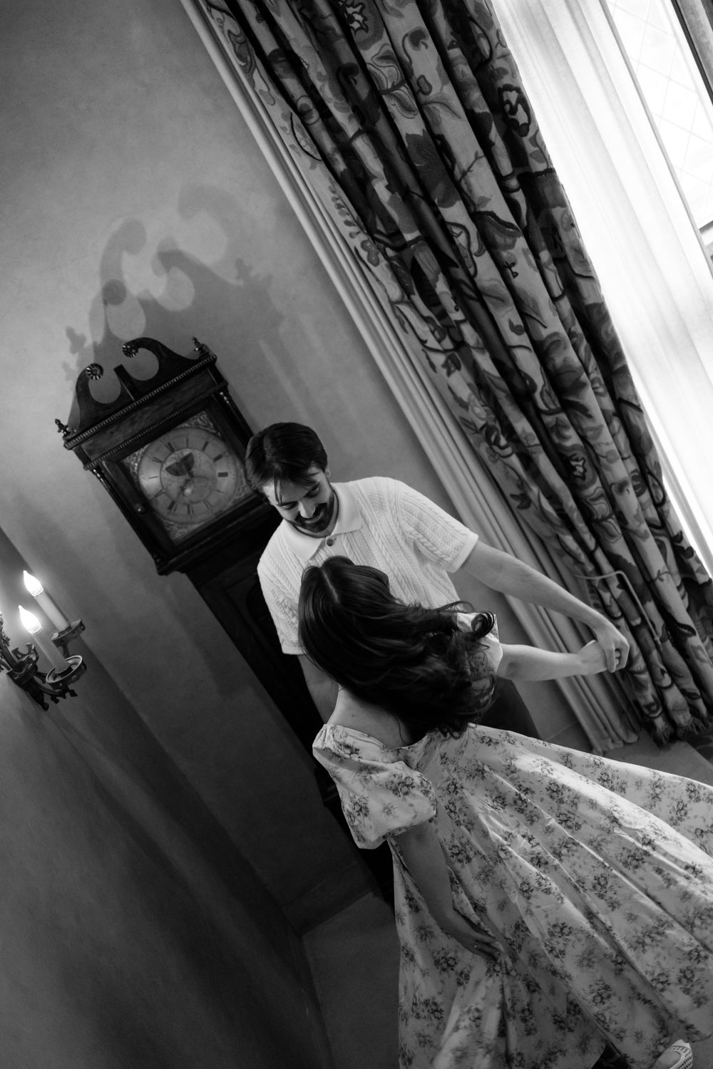 Black and white photo of a couple dancing together near a grandfather clock in an elegant room.