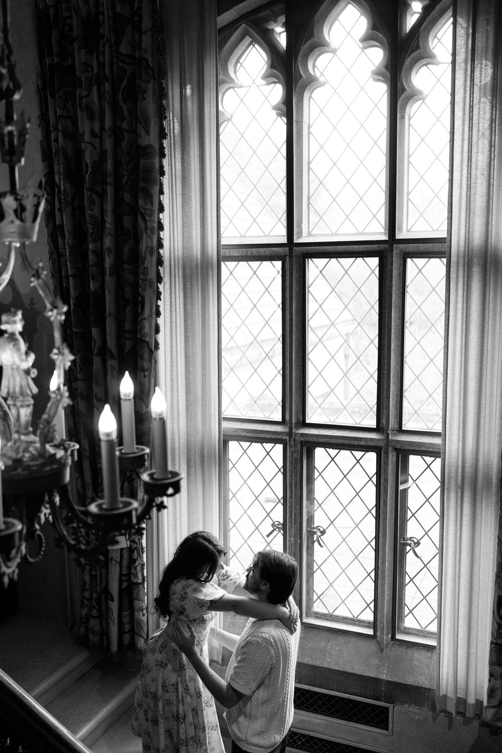 Black and white photo of couple embracing by tall gothic leaded glass windows with candelabra nearby.