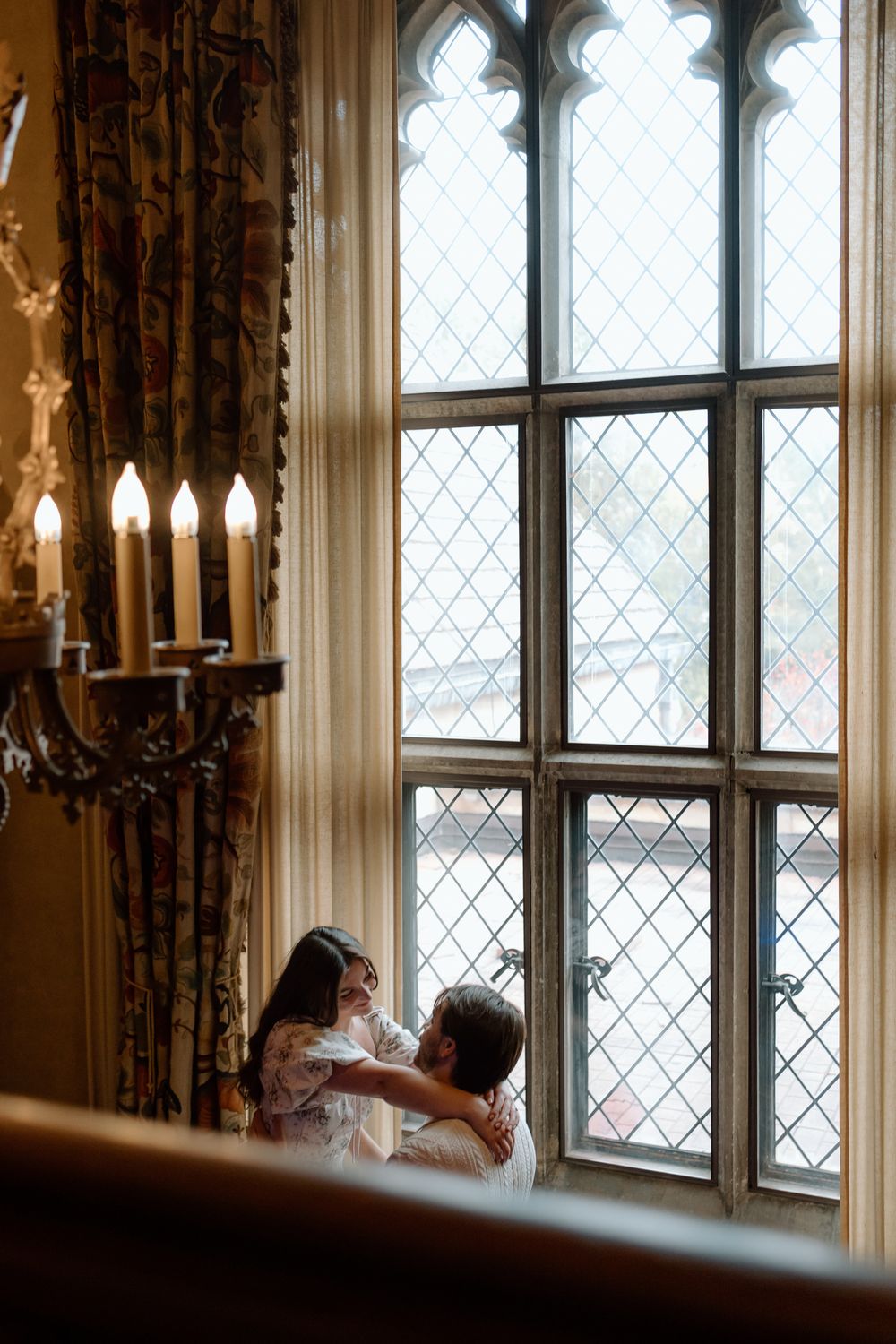 Couple embracing by a grand leaded glass window in an elegant historic venue with candlelit wall sconces.