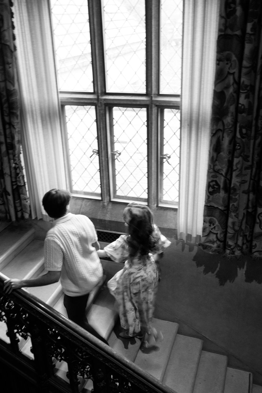 Black and white aerial view of a couple standing on stairs looking out a large latticed window.
