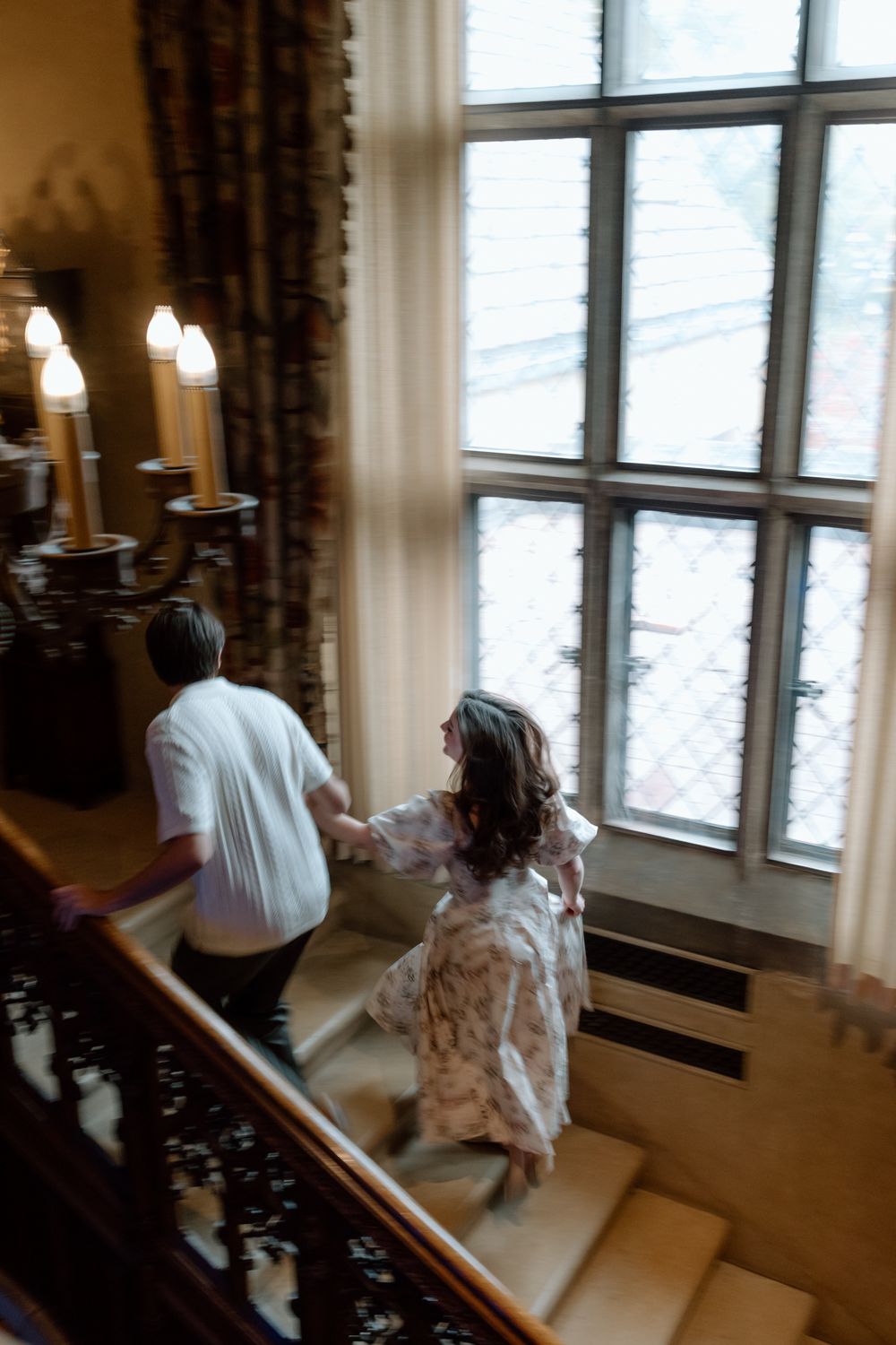 Couple holding hands while ascending a grand staircase past candelabras and large windows in a manor.
