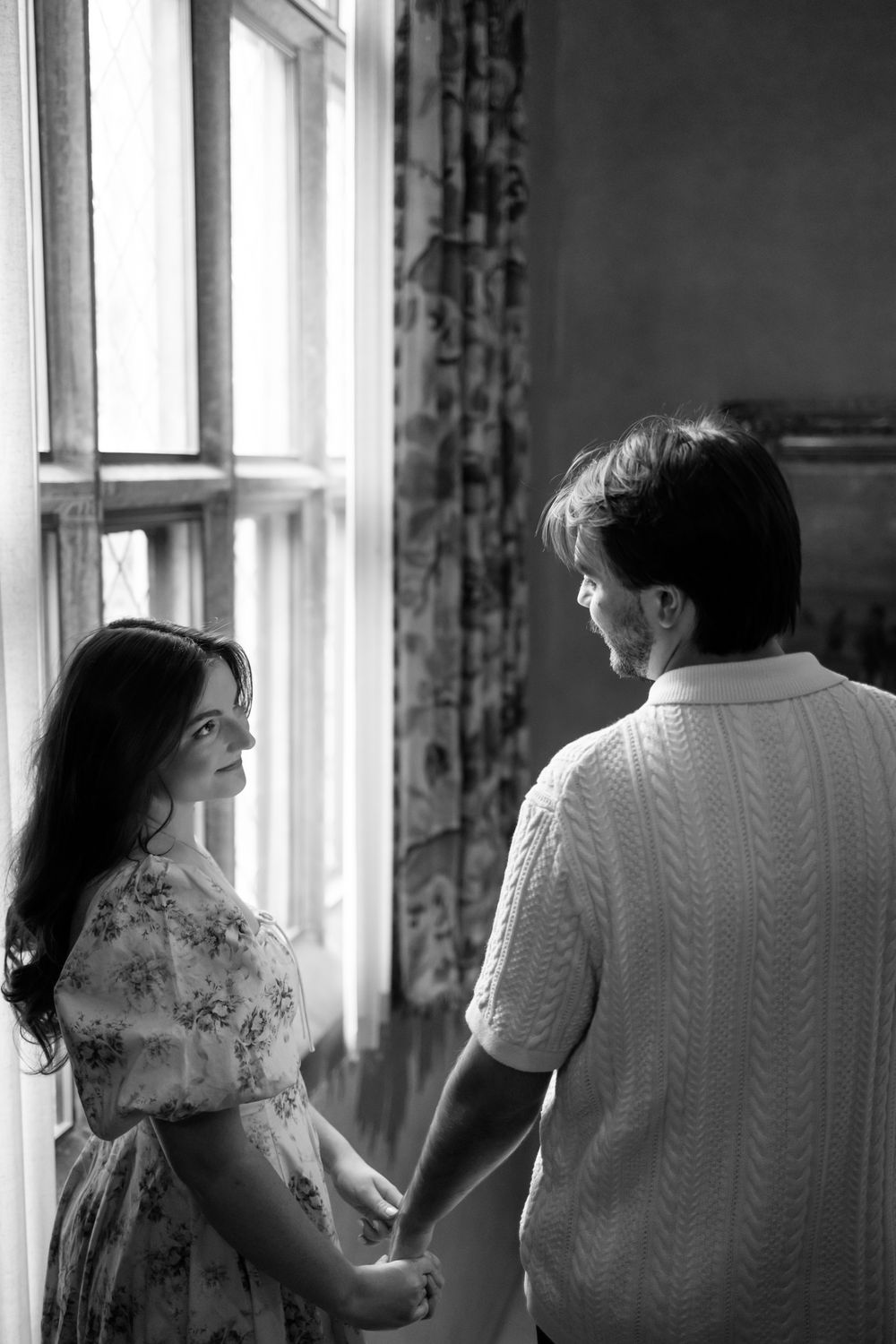 Black and white photo of a couple holding hands by a sunlit window with floral curtains.