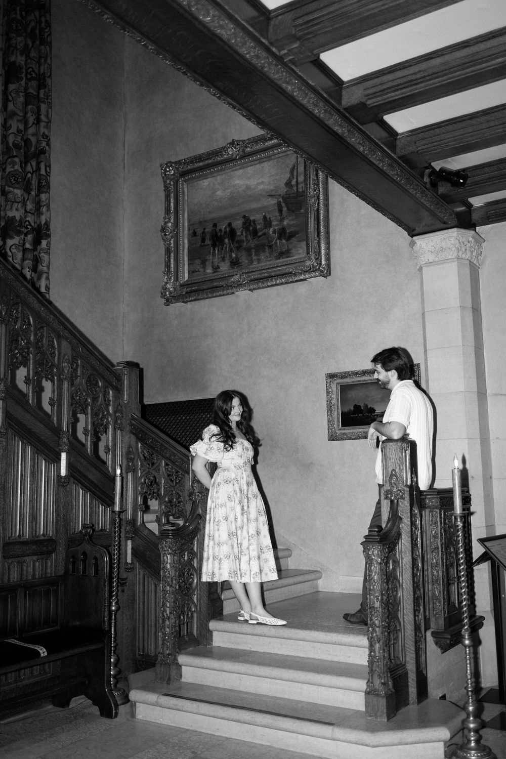 Black and white photo of a couple on ornate wooden stairs in a grand historic mansion interior.