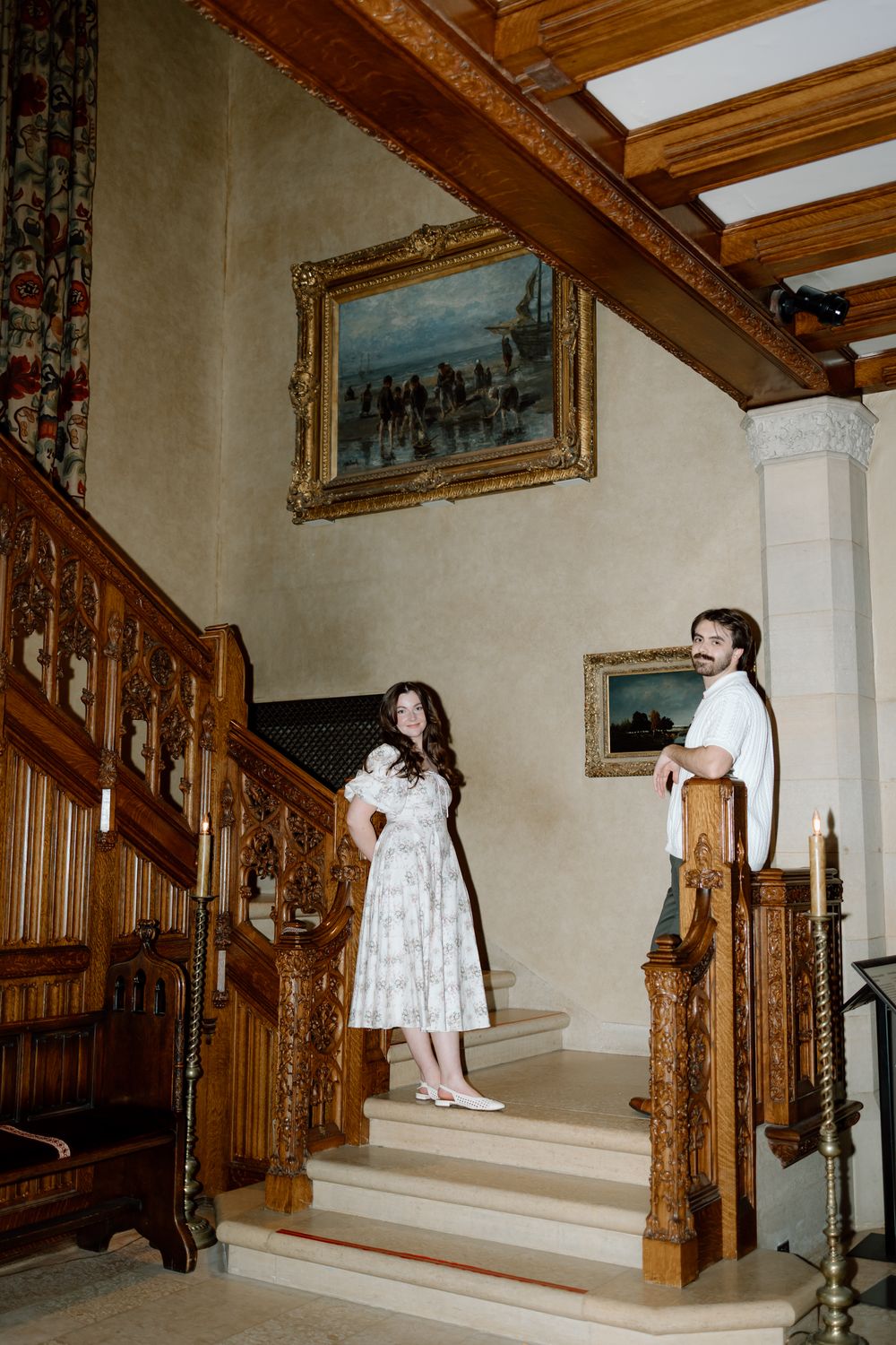 Couple standing on ornate wooden staircase in elegant manor with classic oil paintings.