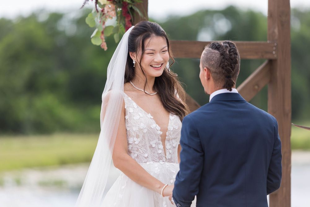Radiant bride in floral gown laughs with groom during outdoor lakeside wedding ceremony.