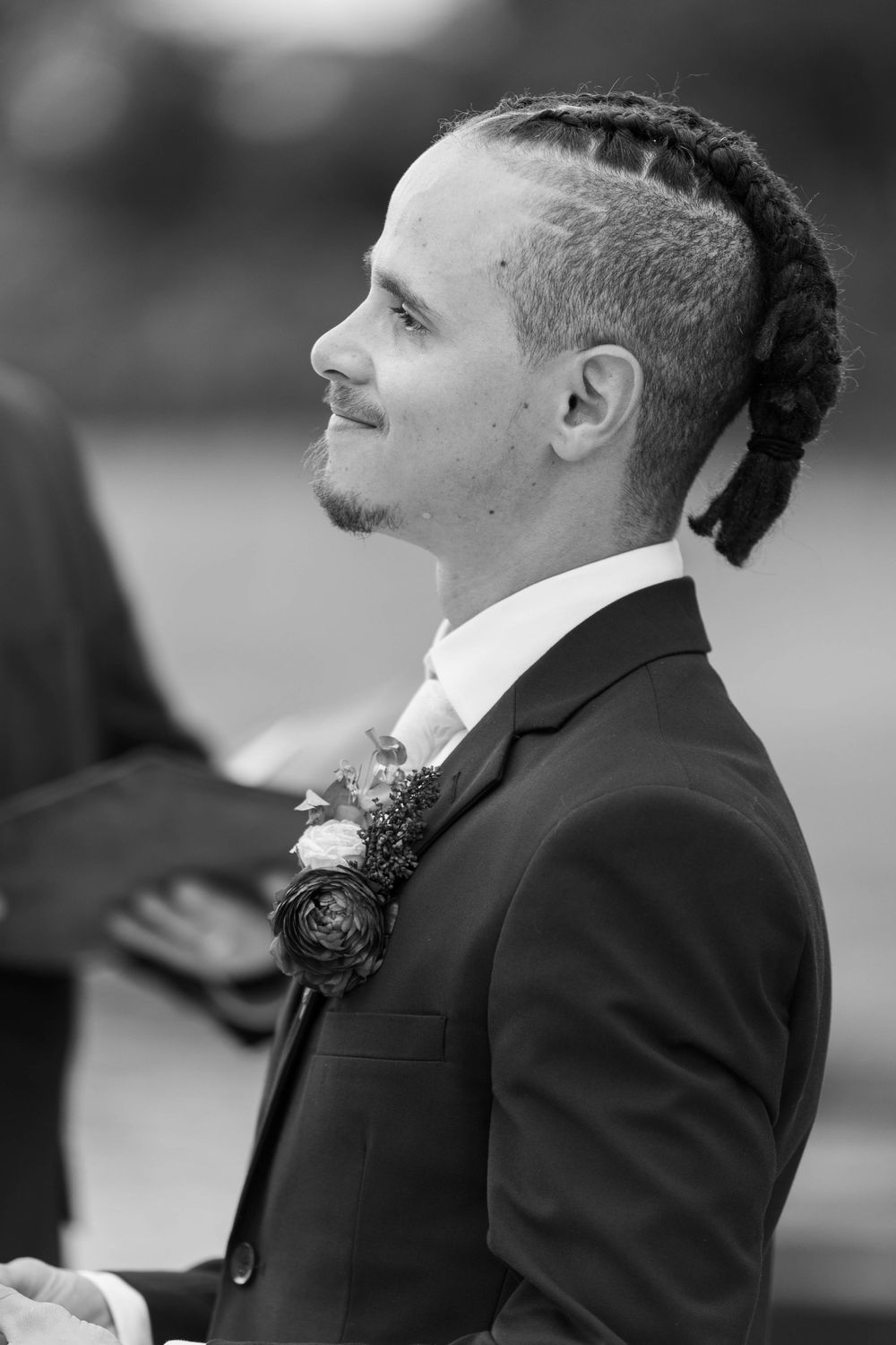 Black and white portrait of a groom in a suit with boutonniere and mohawk hairstyle at a wedding ceremony.