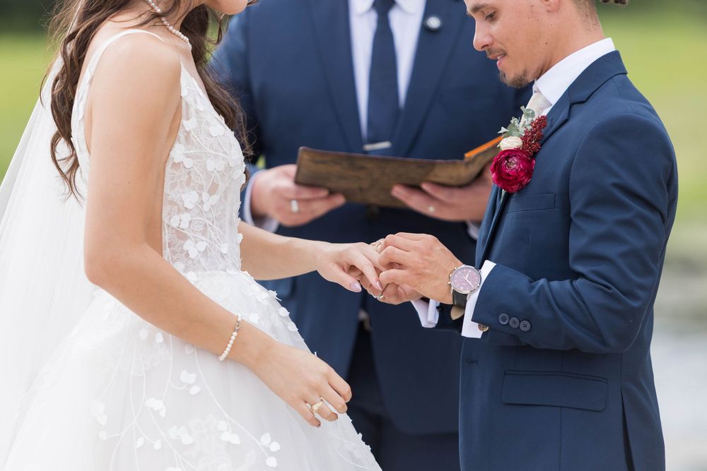 Groom places wedding ring on brides finger during outdoor ceremony.