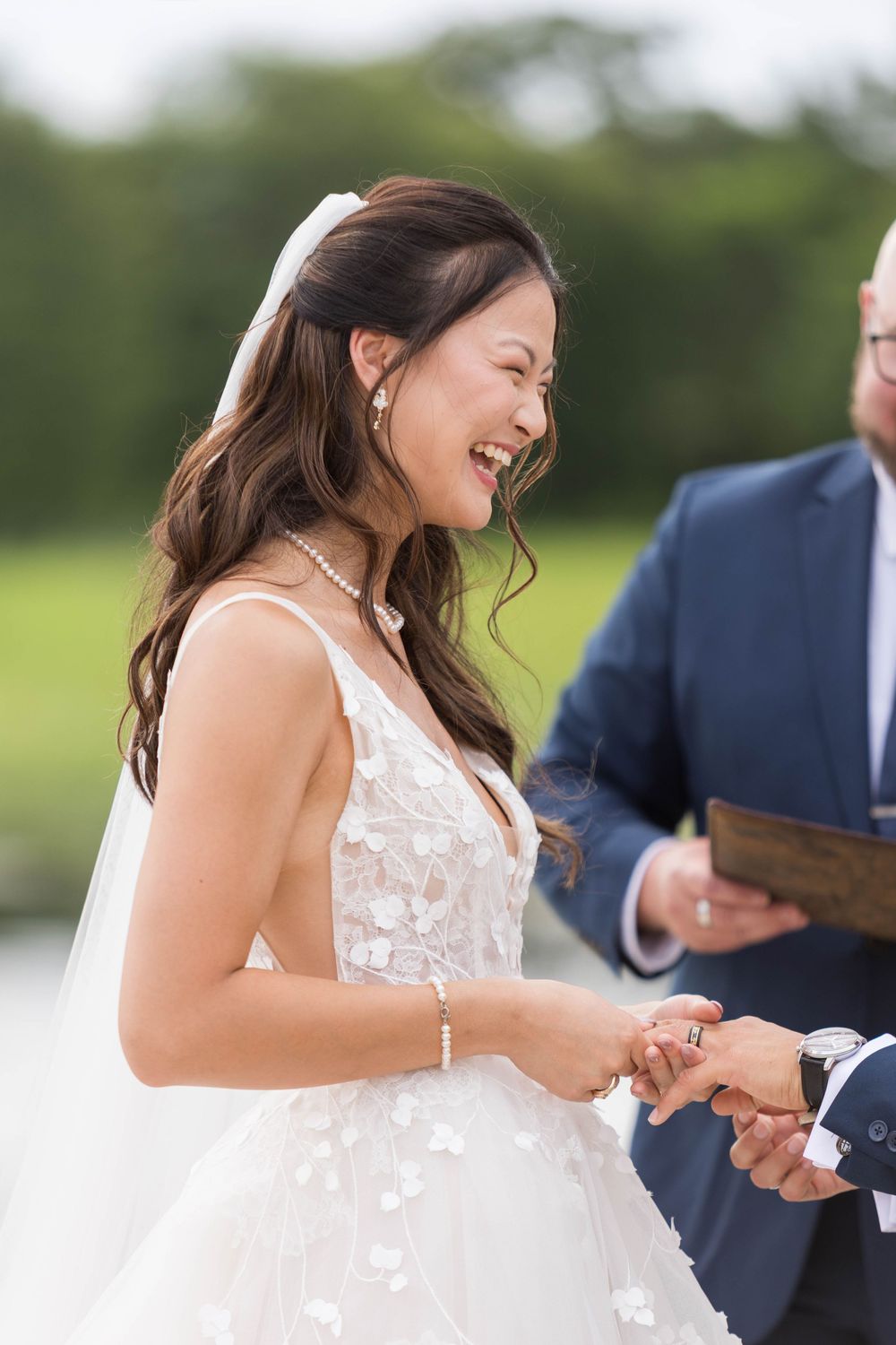Bride laughs joyfully during ring exchange at outdoor wedding ceremony by the water.