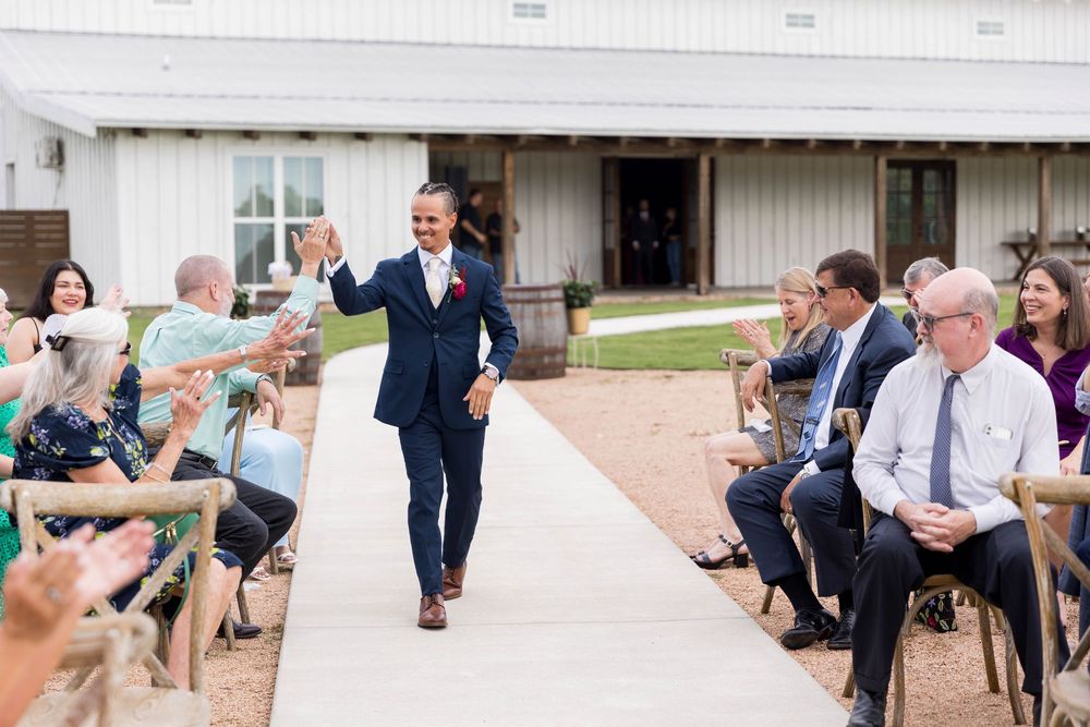 Groom walks down the aisle in navy suit, smiling and waving at wedding guests seated outdoors near a white barn.