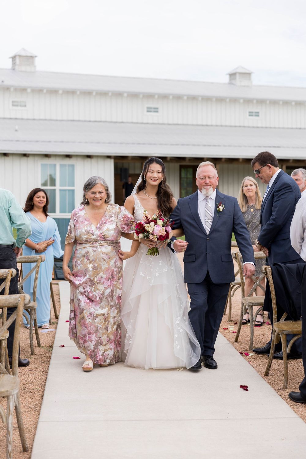 Bride walks down the aisle with parents at outdoor barn wedding ceremony.