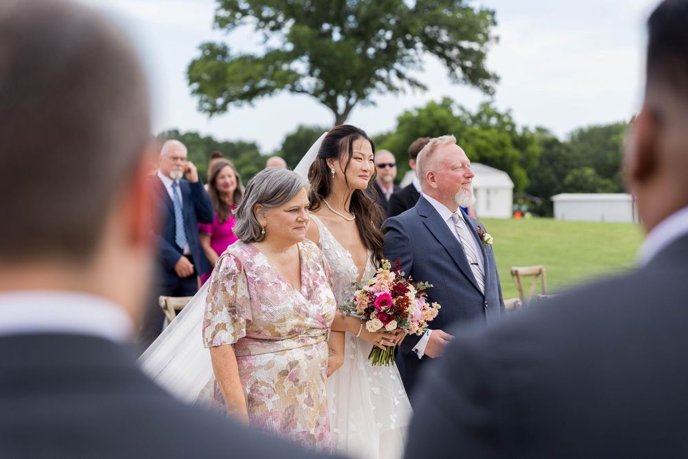 Bride walks down outdoor aisle with parents, holding floral bouquet, guests seated on green lawn at elegant wedding ceremony.