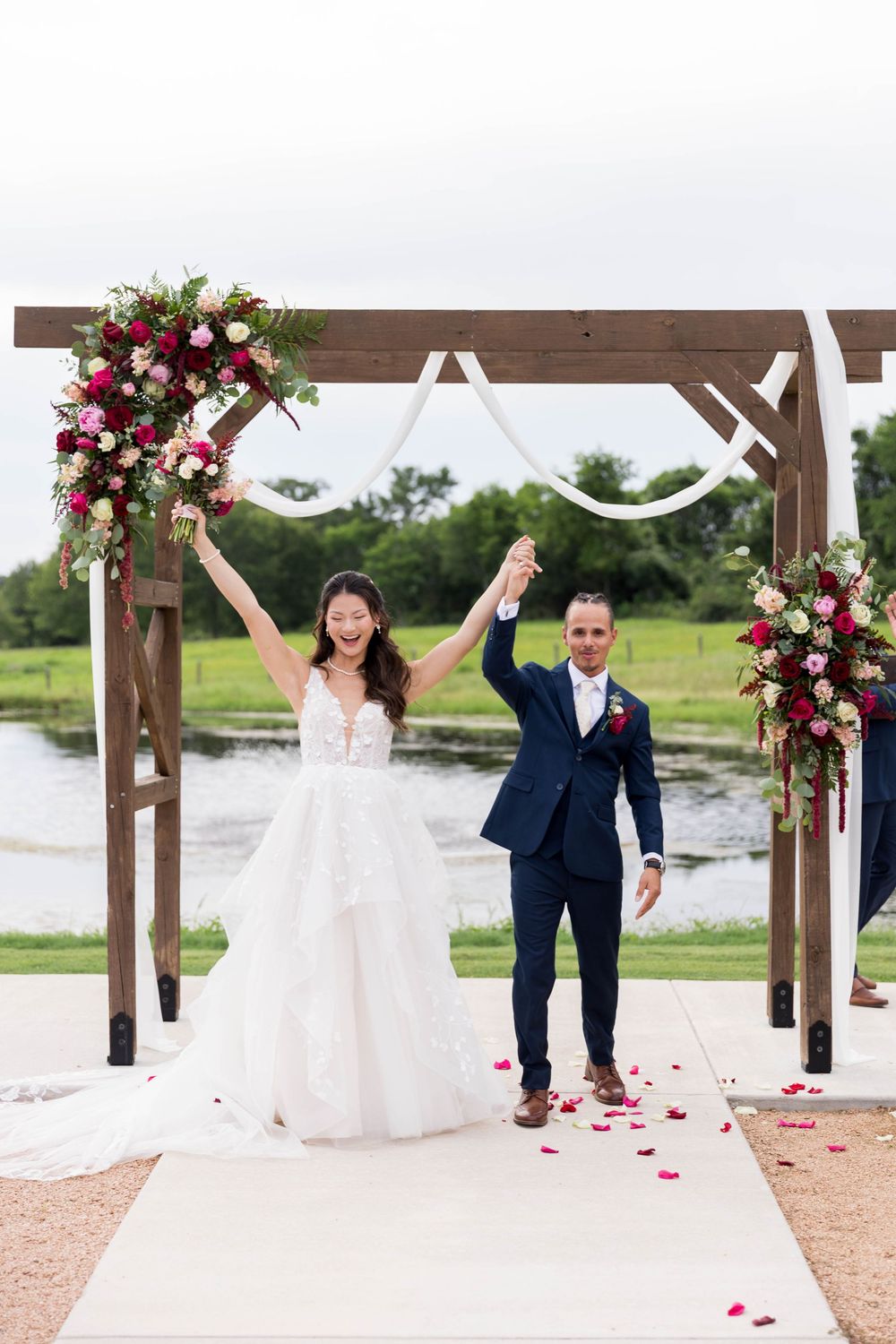 Joyful bride and groom raise hands under floral arch at outdoor waterfront wedding ceremony.