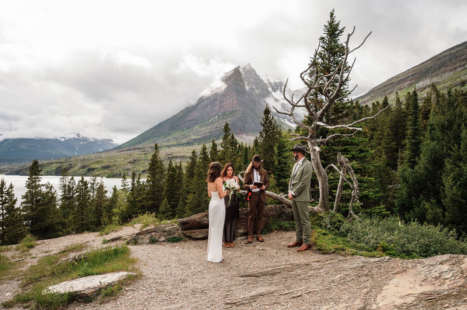 Intimate mountain elopement ceremony with couple and officiant surrounded by alpine trees and dramatic rocky peaks.
