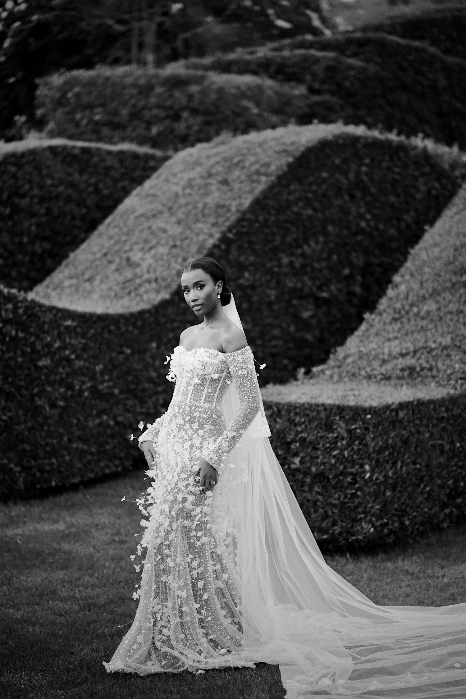 Black and white portrait of bride in floral applique off-shoulder gown with long train before sculpted topiary hedges.