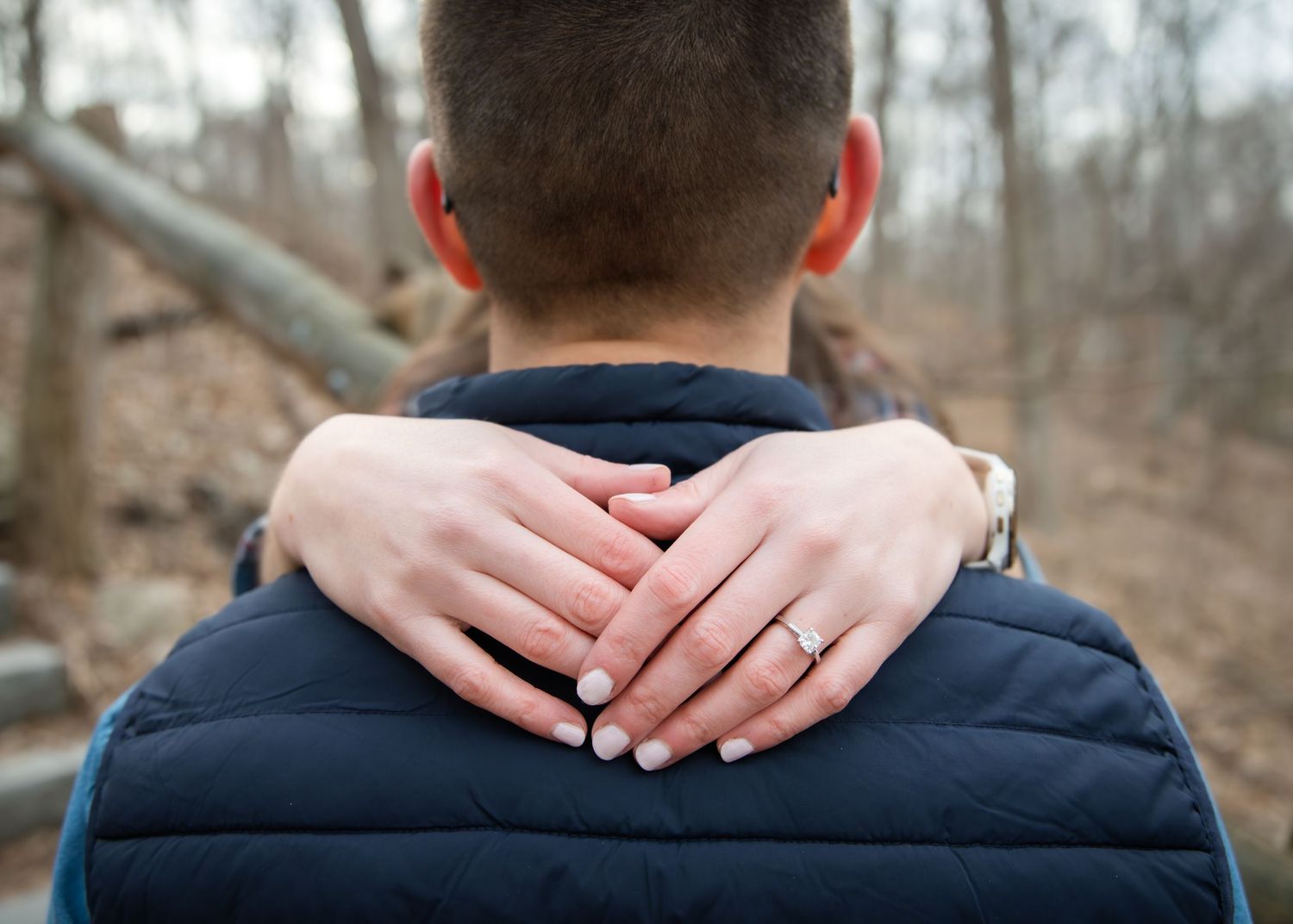 Woman's hands with engagement ring resting on man's shoulders outdoors in wooded setting.